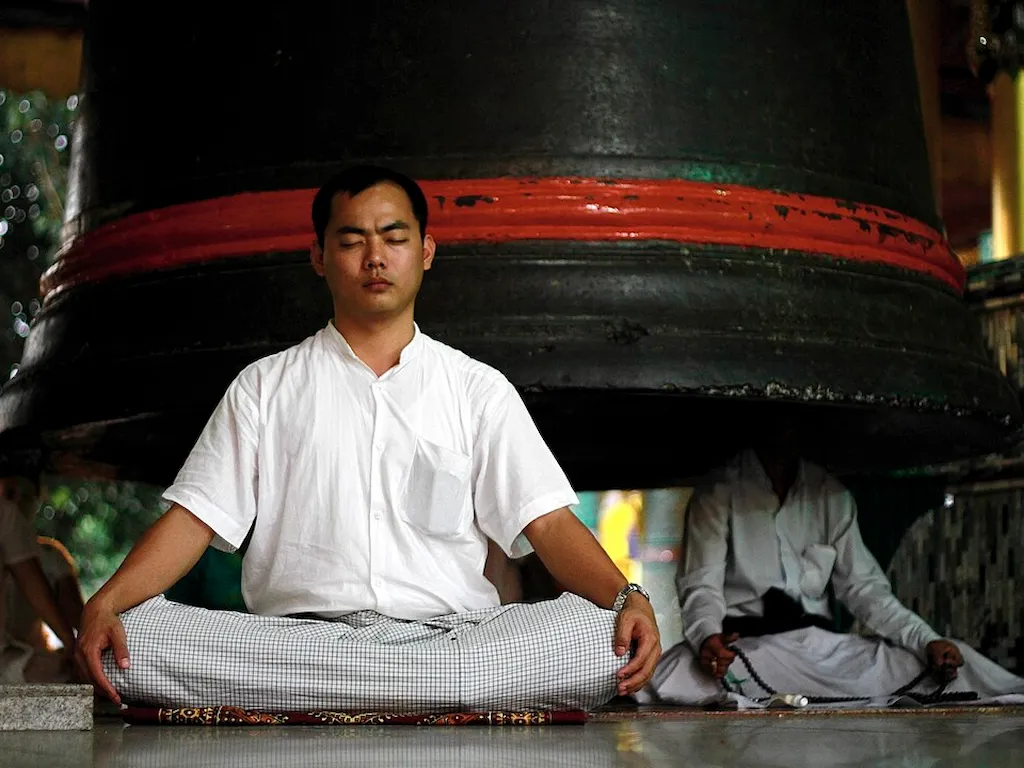 Büyük bir çan önünde meditasyon yapan Budist rahip, Budizm etik mi yoksa aydınlanma mı sorusunu yansıtan ruhsal uygulama sahnesi.